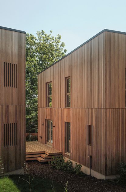 Two modern wooden buildings with vertical slat siding flank a wooden deck courtyard, with trees and a clear blue sky behind.