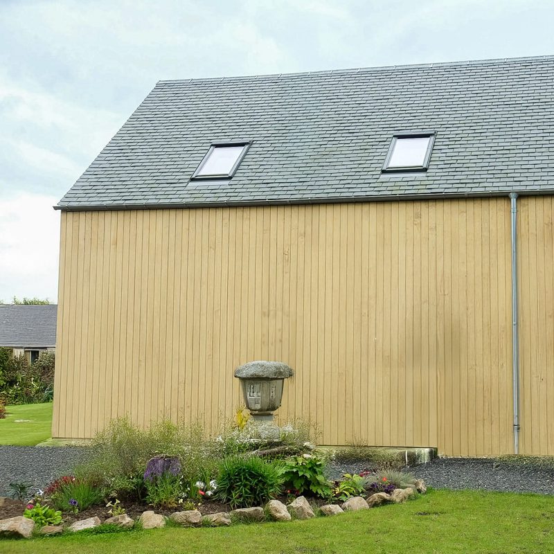 Beige wooden building with a slate roof and two skylights; a stone pedestal garden ornament sits in a rock-bordered flower bed by a gravel path.