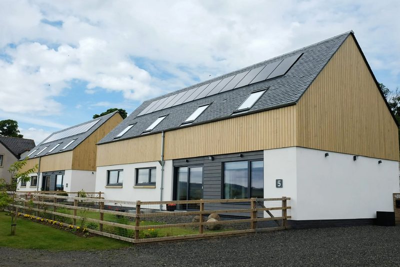 Row of modern semi-detached homes with white lower walls and wood-clad upper facades, gray roofs with solar panels; fenced front gardens.