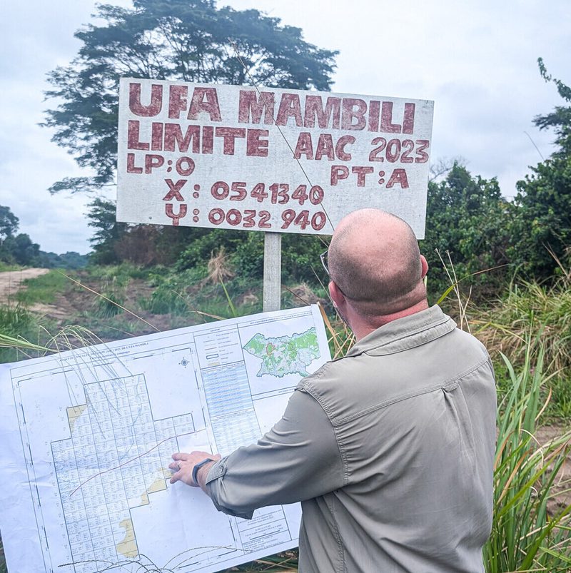 Back view of a bald person in a light gray shirt examining a large map outdoors, with a weathered red-letter sign and tall grasses behind.