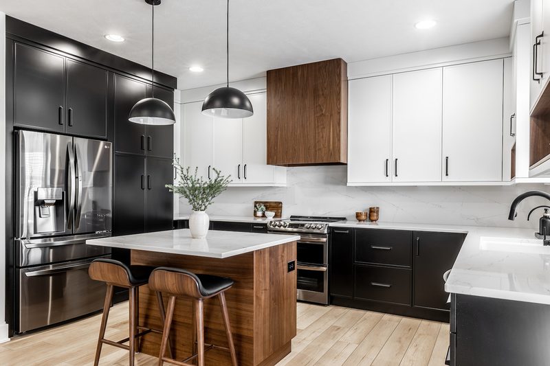 Modern kitchen with black lower cabinets and white upper cabinets, fridge, and a wood-accent island with a white countertop. Pendant lights, stools, marble countertops.