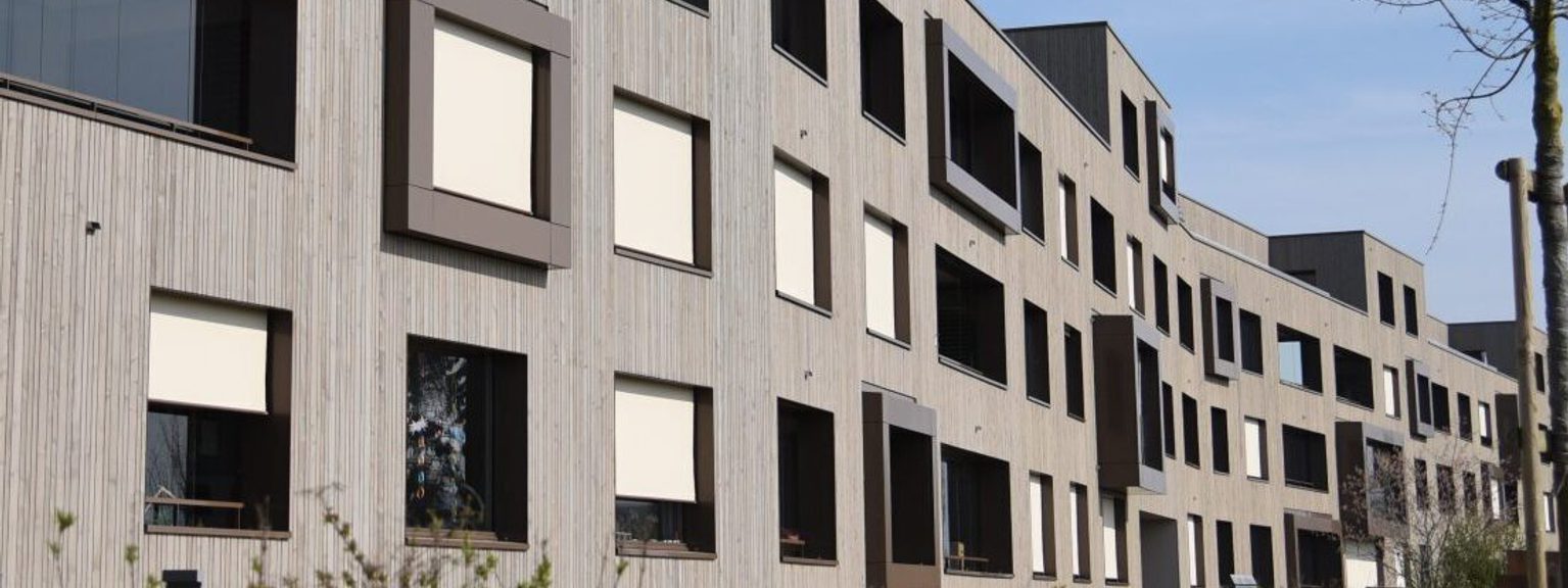 Modern multi-story apartment building with vertical wood siding, windows with shading panels, along a street under a clear sky and a leafless tree.