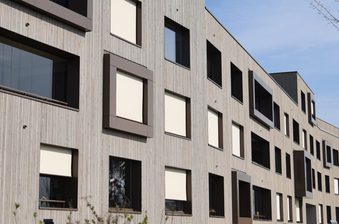 Modern multi-story apartment building with vertical wood siding, windows with shading panels, along a street under a clear sky and a leafless tree.