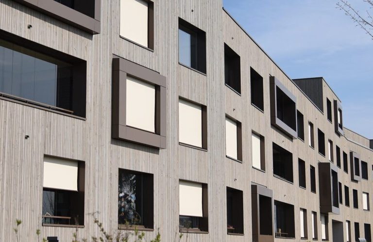 Modern multi-story apartment building with vertical wood siding, windows with shading panels, along a street under a clear sky and a leafless tree.