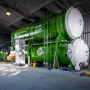 Inside a metal warehouse, a large green cylindrical machine labeled BURN BLOCK and Timber Treatments, with white end caps and visible pipes.