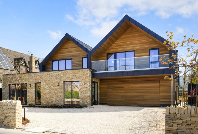 Modern two-story home with stone ground floor, wood upper level, blue-trim gable roof, glass balcony, and tall wooden garage door; gravel driveway.