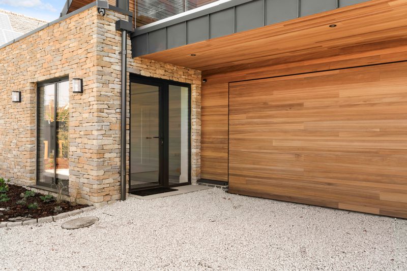 Modern house exterior with a stone facade, a wooden garage door, a glass entry door, a gravel driveway, and a small garden bed with wall lights.