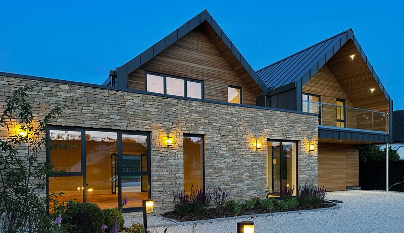 Contemporary two-story home with stone facade, wooden upper siding, and two gabled roofs. Glass doors and warm lights glow over a gravel driveway with lavender.