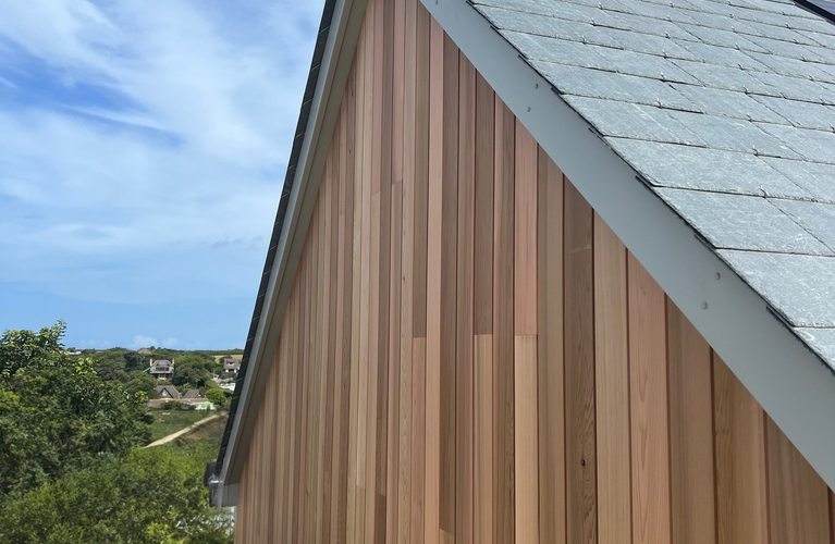 Vertical wooden siding on a house with a steep gray shingle roof, white trim, and a red ridge cap, set against blue skies and distant trees.