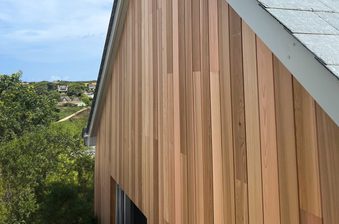 Side of a building clad in vertical wooden planks in brown tones, with a gray shingled roof; greenery and distant houses beneath a blue sky.
