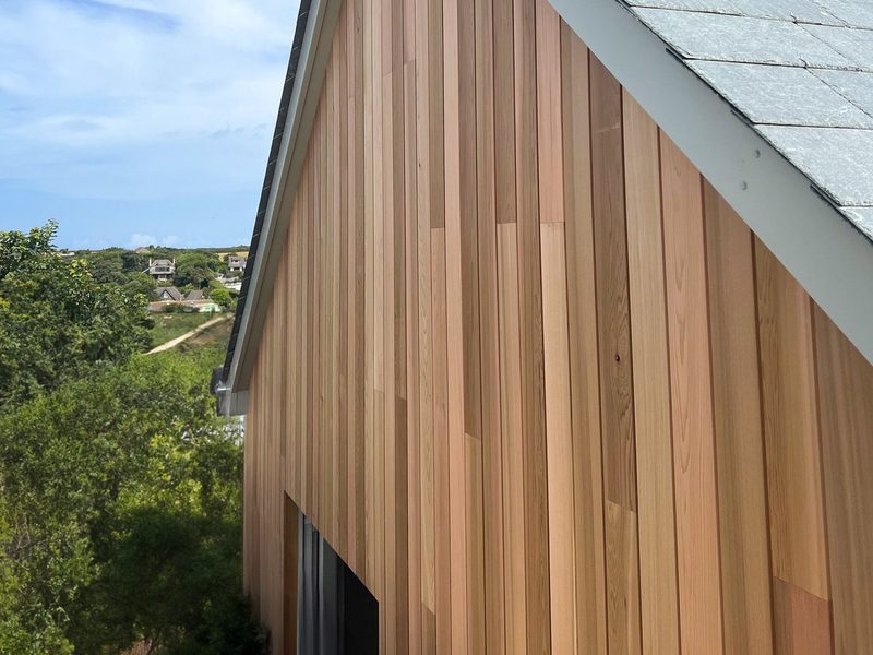 Side of a building clad in vertical wooden planks in brown tones, with a gray shingled roof; greenery and distant houses beneath a blue sky.