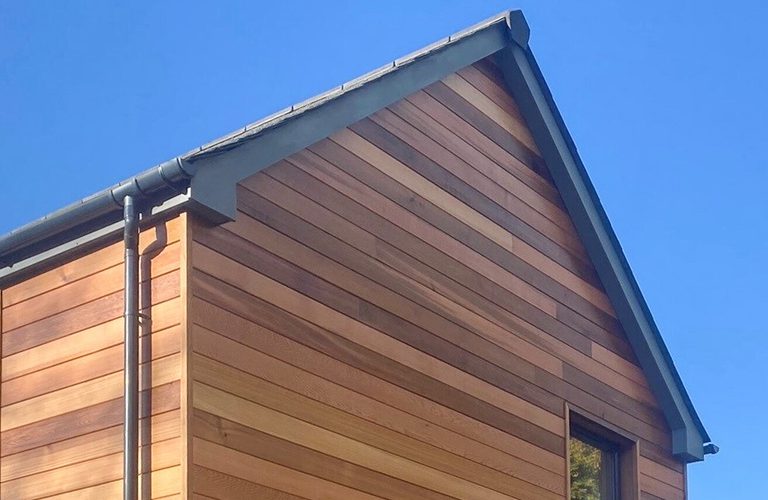 Close-up of a wooden house exterior with horizontal planks, a sloped roof, gray trim, a vertical gutter on the left, and a window against a blue sky.