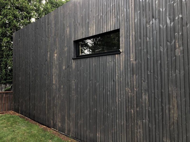 Dark vertical wooden wall with a long narrow horizontal window; a green lawn and trees on the left, with a wooden fence in the background.