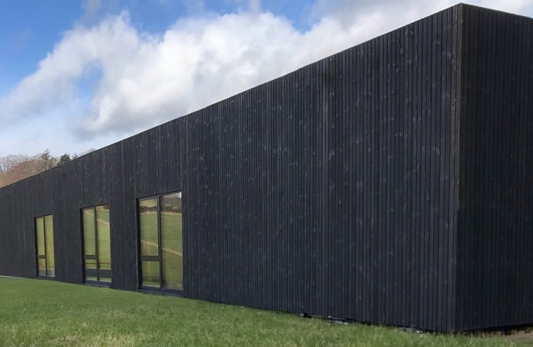 Long, dark wooden building with vertical slats and large glass doors sits on a grassy field beneath a blue, partly cloudy sky.