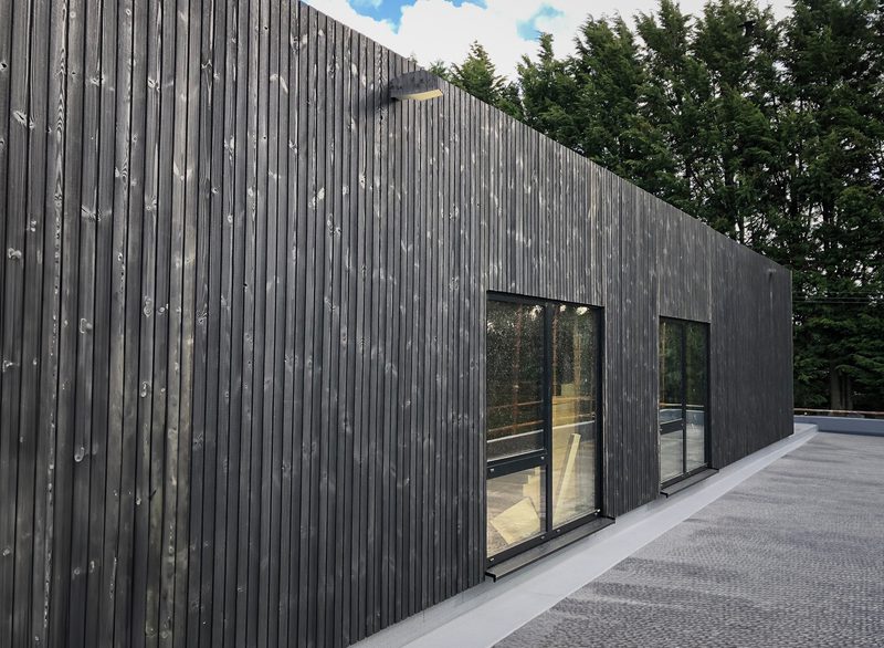 Modern dark wood-clad building with tall vertical panels, two large glass doors, and a flat gray deck, set beside evergreen trees under a blue sky.
