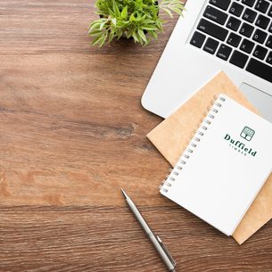 Top-down view of a wooden desk with a laptop, a small green plant, a white spiral notebook labeled Duffield Timber, an envelope, and a silver pen.