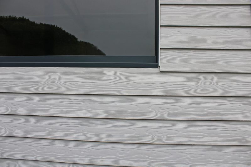Close-up of light gray wooden clapboard siding with visible grain, and a dark-framed window near the top left.