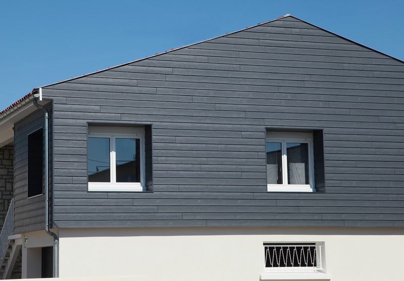 Two-story house with dark blue-gray horizontal siding, two white-framed upper windows, a white lower facade, and a steep gable roof under a clear blue sky.
