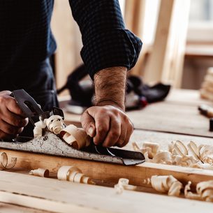 A person in a dark plaid shirt uses a hand plane to shave wood on a workbench, with wood shavings scattered around.