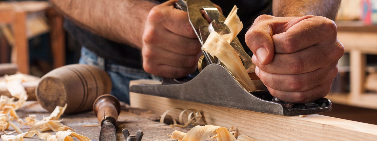 Close-up of hands planing a wooden board with a hand plane; shavings and carving tools litter a workshop bench.