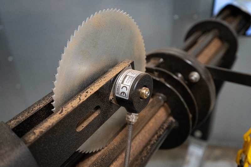 Circular saw blade on a dusty metalworking machine, with a small cylindrical sensor and cable; a belt-driven shaft visible in the background.