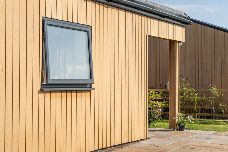 Tan vertical wooden siding on a modern building, with a gray-framed window on the left and an open doorway on the right, a stone path and small garden beyond.