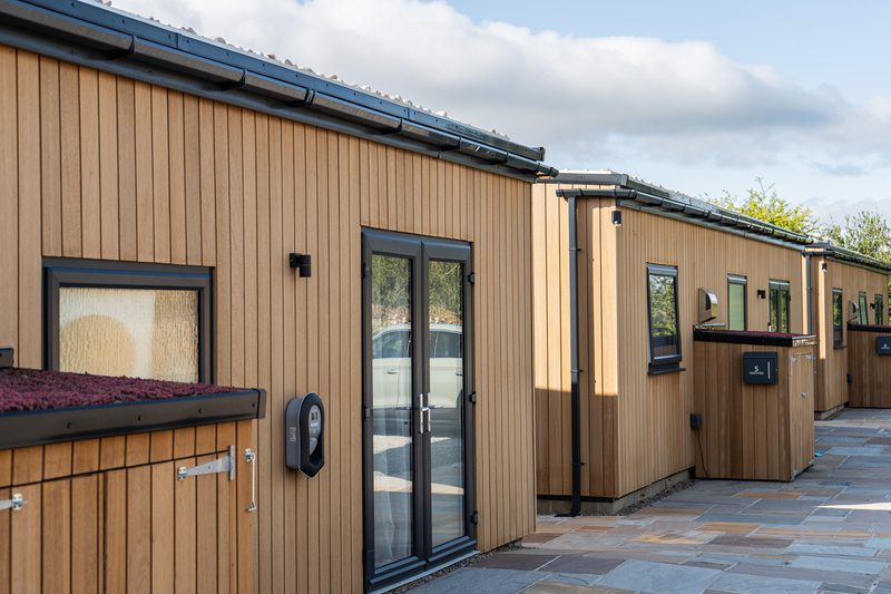 Row of light-brown wooden houses with black trim and glass doors along a paved walkway under a blue sky with clouds.