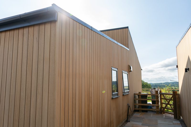 Brown wooden-clad building with vertical panels, two small windows, sun glare, and a wooden gate on a stone patio overlooking rolling countryside.
