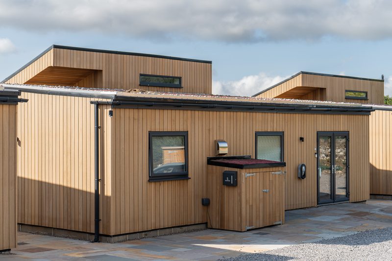 Modern wooden building with vertical tan cladding, flat roofs, glass doors, a mailbox, and a cabinet-like outdoor unit; blue sky above.