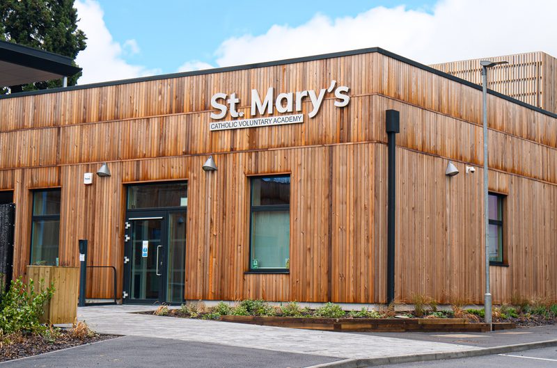 Wooden-clad school building with a "St. Mary's Catholic Voluntary Academy" sign, glass doors, windows, small landscaped beds, and a blue sky above.