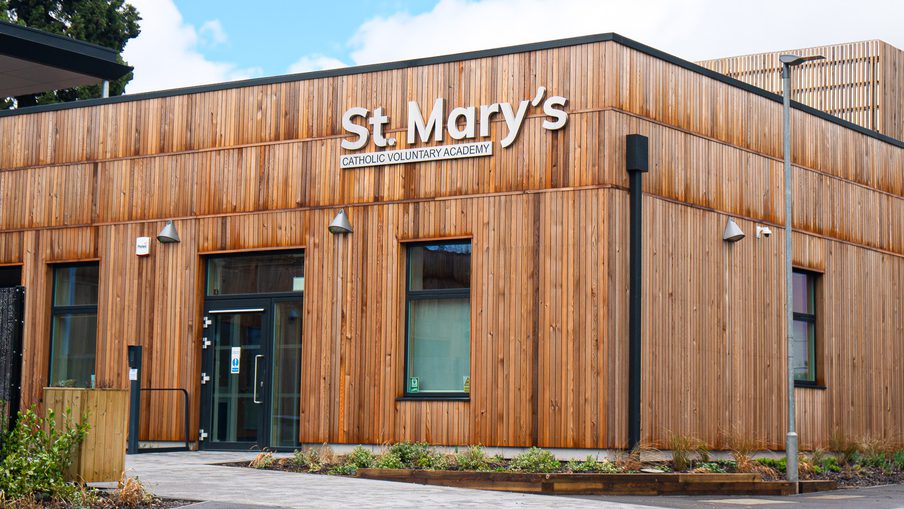 Wooden-clad school building with a "St. Mary's Catholic Voluntary Academy" sign, glass doors, windows, small landscaped beds, and a blue sky above.