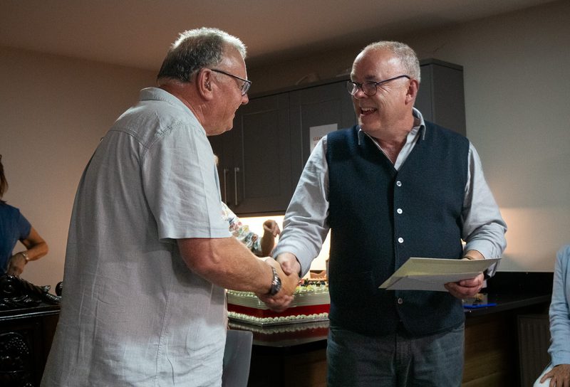 Two adults shake hands; one in a light short-sleeve shirt and glasses, the other in a dark vest with a certificate, cake in background.