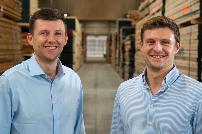 Two individuals in light blue shirts stand in a warehouse aisle flanked by stacked lumber, smiling at the camera.