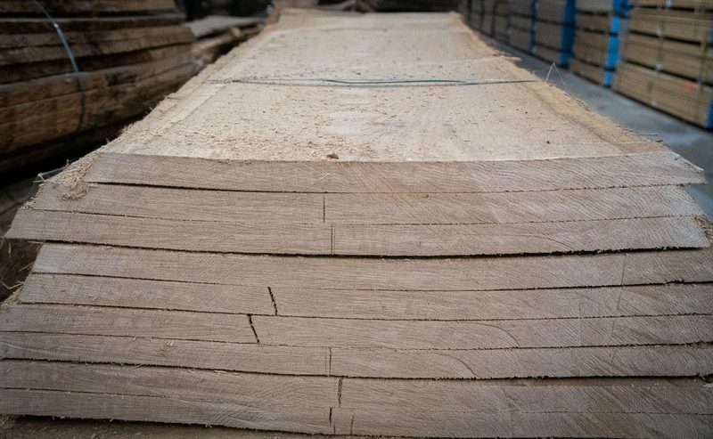 Stack of rough-sawn wooden boards in a lumberyard, pale brown with visible grain and cracks, extending into the background.
