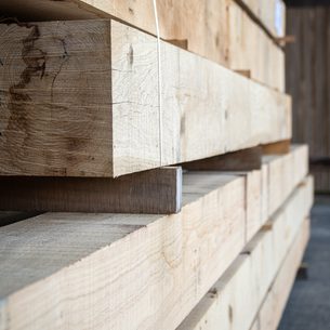Stack of rough wooden boards in a lumber yard, bound with a white strap; sunlight highlights the grain across the planks.