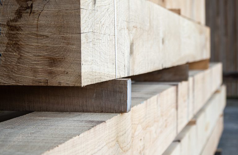 Stack of rough wooden boards in a lumber yard, bound with a white strap; sunlight highlights the grain across the planks.