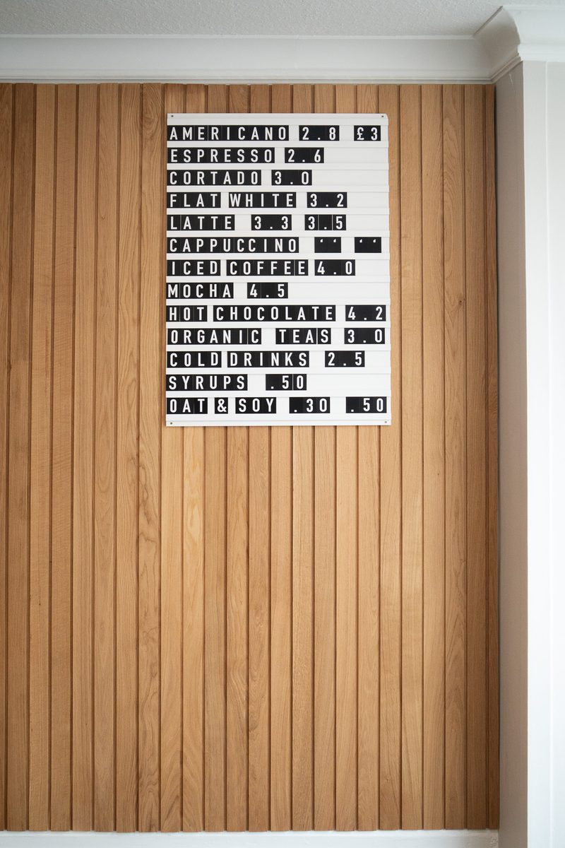 Letterboard menu mounted on a vertical wood-paneled wall listing coffee drinks and prices (Americano, Espresso, Latte, Cappuccino, iced coffee).