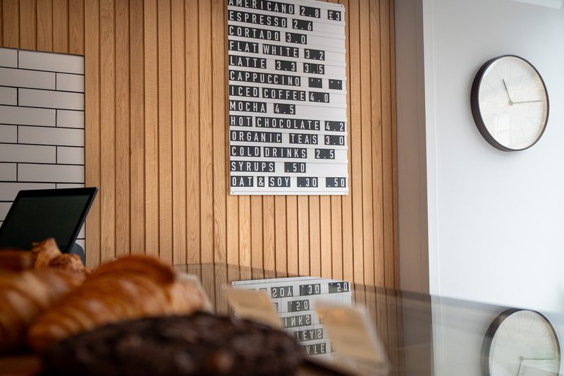 Cafe interior with a wooden slat wall, a drink menu board, a round clock on the white wall, croissants on a glass counter, and a laptop on the left.