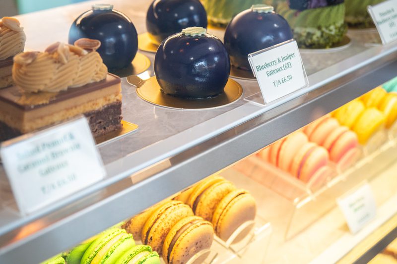 Assorted pastries in a glass display: glossy dark blue mirror-glazed domes on gold bases, a layered cake, and rows of colorful macarons.