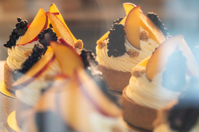 Cupcakes with white swirled frosting topped with peach slices, blackberries and crumbs, arranged in a bakery display case.