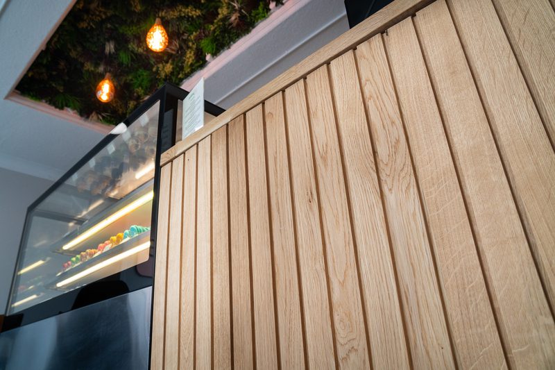Wood slat wall beside a glass pastry display with colorful sweets; overhead greenery and warm orange pendant lights.