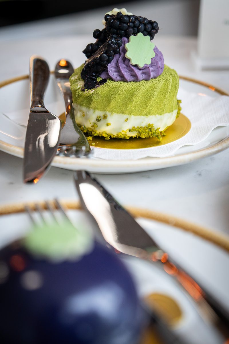 Green matcha cake with a white cream layer and pistachio crumb base, topped with purple frosting, blackberries, and a green leaf on a gold-rimmed plate with silverware.