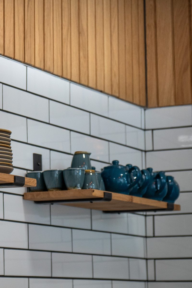 Kitchen wall with white subway tiles and wooden paneling, two floating shelves display blue teapots and mugs, with a stack of plates on the left.