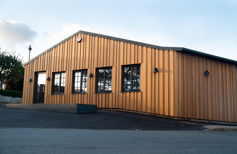 Single-story building with vertical wooden siding, orange-brown panels, several rectangular windows, a door, exterior lights on a paved lot.