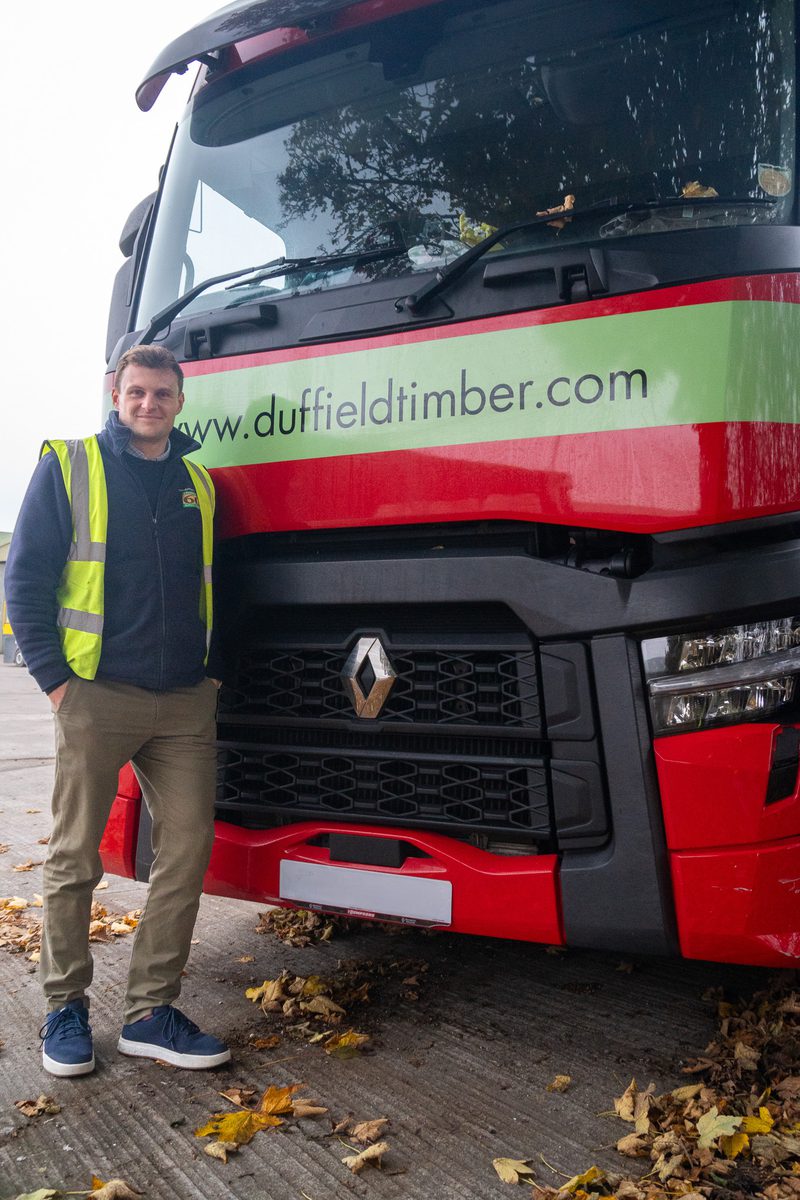 Person in a high-visibility vest stands beside a large red Renault truck with a lime-green stripe displaying duffieldtimber.com; autumn leaves on the pavement.