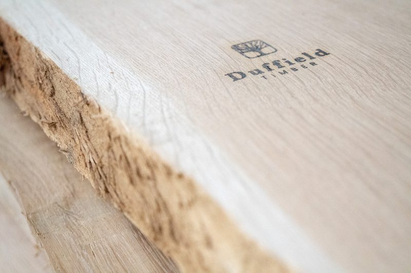 Close-up of light wood surface with a rough left edge; stamped logo reads 'Duffield Timber' with a shield icon.