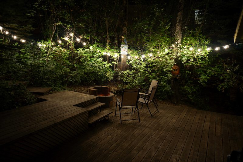 Nighttime backyard wooden deck with string lights, a circular fire pit, two chairs, a wooden bench/steps, and dense green foliage in the background.