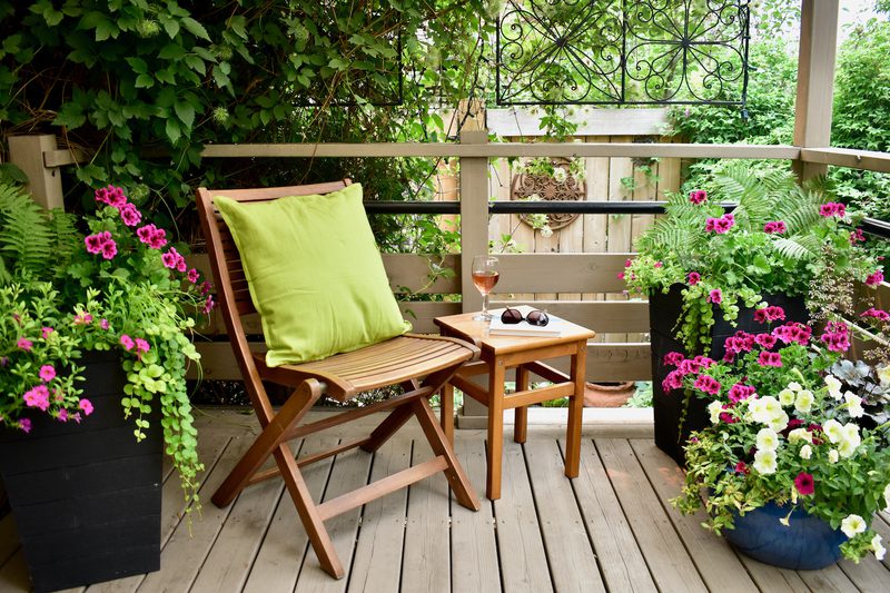 Cozy wooden balcony on a deck with a lime-cushioned chair, a small table holding sunglasses and a wine glass, flanked by vibrant pink, white, and green flowers in planters.