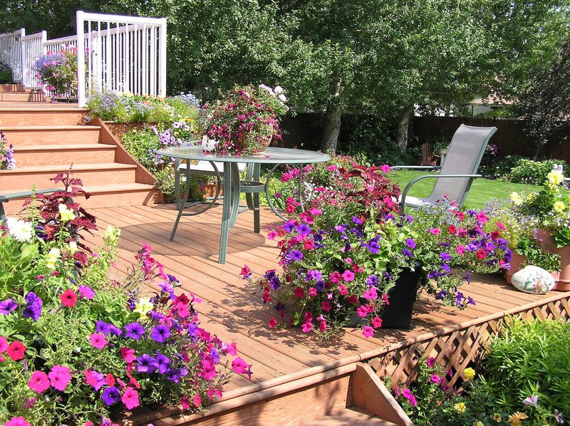 Wood deck with stairs, colorful flowers in pots, a round glass table with a floral centerpiece, and a gray lounge chair amid a lush garden.
