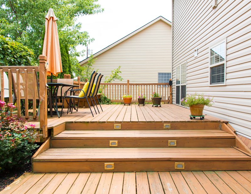 Wooden deck with steps, patio table and chairs under an orange umbrella, and potted flowers along the house in a fenced backyard.